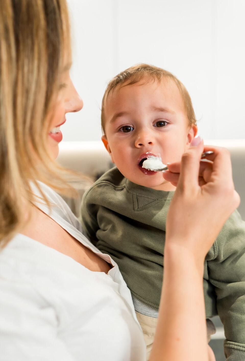 Mother feeding her baby son with yogurt in a spoon in the kitchen
