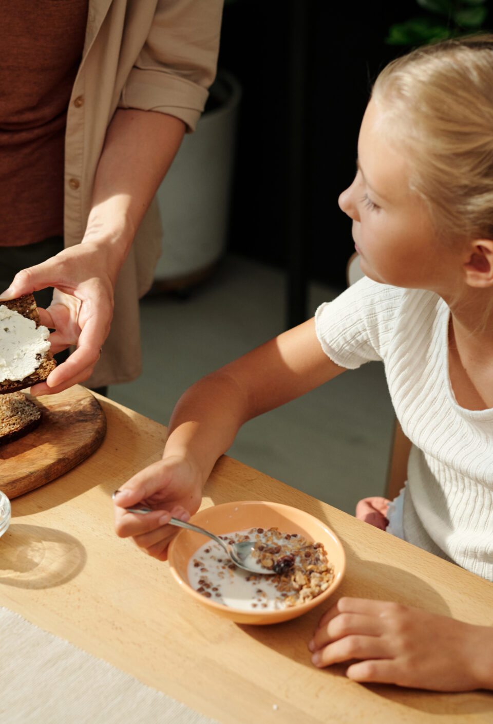 Youthful girl eating muesli with milk and looking at her mother making sandwich